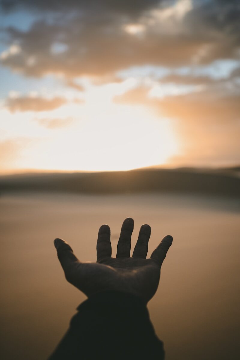 A hand reaching toward a bright low sun in a warm hazy sky